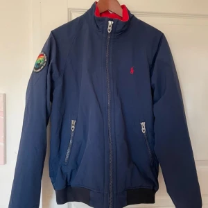 Blå jacka Polo Ralph Lauren M - Snygg marinblå vindjacka från Polo Ralph Lauren med röd fleecefoder, dragkedja framtill och två fickor med dragkedja. Jackan har en patch på ena ärmen och klassisk röd logga på bröstet. Perfekt för kyliga dagar och stilren streetlook.