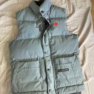 Grå dunväst från Canada Goose XS - Säljer en grå dunväst från Canada Goose med hög krage, stora fickor framtill och broderad röd lönnlöv-detalj på bröstet. Västen har tryckknappar och dragkedja samt klassisk Canada Goose-logga på insidan. Perfekt för lager-på-lager under kyliga dagar.