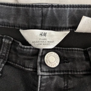 Svarta flare jeans från H&M - Säljer ett par svarta/mörkgråa flare jeans från H&M med låg midja och justerbar midja. Jeansen har klassisk design och är tillverkade i mjukt denimtyg. 