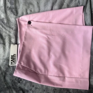 Rosa omlottkjol från Karl Lagerfeld - Supersnygg rosa omlottkjol från Karl Lagerfeld med stilren design och en diskret knappdetalj i midjan. Kjolen är kort och har en clean look utan mönster, perfekt för dig som gillar en chic vibe. Materialmix av polyester, ull och viskos för en lyxig känsla.