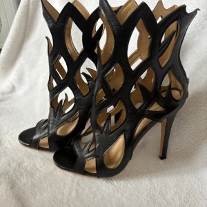 Svarta cut-out pumps med hög klack. Stl.37 - Unika svarta pumps med coolt cut-out mönster och hög klack. Skorna har öppen tå och dragkedja bak för enkel påtagning. Tillverkade i skinn med en edgy design som verkligen sticker ut. Perfekta för dig som vill ha något extra till outfiten. (23,5 innermått)