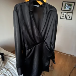 Svart omlottklänning från Zara - Snygg svart omlottklänning från Zara i glansigt material. Klänningen har lång ärm, djup v-ringning och en elegant omlott-detalj framtill. Perfekt för dig som gillar stilrena och trendiga plagg med en lyxig känsla. Silkes material 