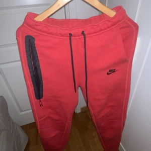 Röda mjukisbyxor från Nike - Säljer ett par röda mjukisbyxor från Nike med svart logga på vänster ben. Byxorna har svarta snören i midjan, en svart dragkedja på ena fickan och ribbade muddar vid bensluten. Perfekt för chill eller träning, riktigt sköna och snygga. Köpta för 1100. använda 2 gånger 