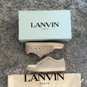 Beige sneakers från Lanvin i mocka - Snygga beige sneakers från Lanvin i mjuk mocka med vita snören och vit tå i blankt material. Klassisk låg modell med rund tå och platt sula. Perfekta för dig som gillar stilrena och exklusiva skor med fräsch vibe.