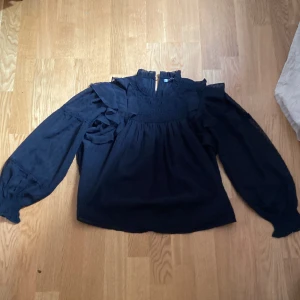 Svart blus med volang från Gina Tricot - Supersnygg svart blus från Gina Tricot med volangdetaljer på axlarna och smock upptill. Långa ärmar med lätt puff och diskret struktur i tyget. Perfekt att styla med jeans eller kjol för en trendig look.