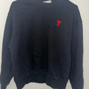 Svart sweatshirt från AMI med rött hjärta - Svart sweatshirt från AMI Alexandre Mattiussi med broderat rött hjärta och A på bröstet. Klassisk rund halsringning, ribbade muddar och avslappnad passform. Perfekt för dig som gillar stilrena plagg med en twist.