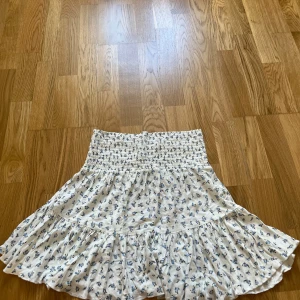 Vit blommig volangkjol med smock - Supersöt kort kjol i vitt med små blå blommor och volangkant. Kjolen har bred smock i midjan som ger en bekväm och snygg passform. 