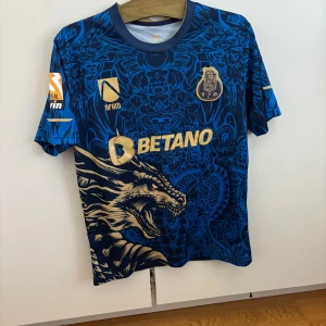FC Porto fotbollströja  - Snygg FC Porto fotbollströja i mörkblått med guldiga detaljer och ett unikt drakmotiv på framsidan. Tröjan har korta ärmar, klubbmärke på bröstet och sponsorer i guld. Tillverkad i lätt och ventilerande material, perfekt för match eller träning eller bara till vardags är den även snygg.