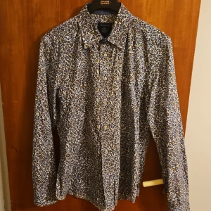 Blommig skjorta från Mel Richman - Snygg skjorta från Mel Richman med blommigt mönster i blått, brunt och vitt. Skjortan har klassisk krage, långa ärmar och knäppning framtill. Perfekt för dig som vill sticka ut med en cool och färgglad stil.