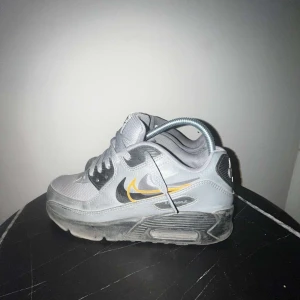 Nike Air Max 90 gråa - Grå Nike Air Max 90 sneakers med svart och gul swoosh på sidan. Ovandel i mesh och syntet, robust sula och klassisk Air Max-dämpning. Skorna har snörning och rund tå, samt tydlig Nike Air-logga på hälen. Perfekta för dig som gillar streetwear och sportig stil.