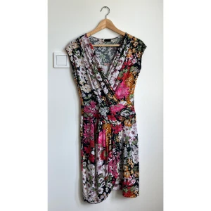 Blommig omlottklänning från Sinéquanone - En omlottklänning från Sinéquanone med färgstarkt blommigt mönster i rosa, vit, grön, svart och orange. Klänningen har v-ringad hals, korta ärmar och markerad midja.