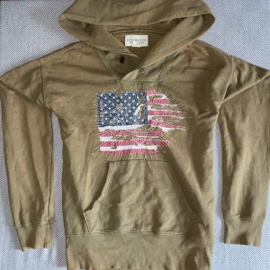 Beige hoodie från Denim & Supply - Säljer en beige hoodie från Denim & Supply Ralph Lauren med coolt tryck av amerikanska flaggan och örn på bröstet. Tröjan har huva, känguruficka och långa ärmar. Perfekt för dig som gillar Grisch och vill sticka ut med en unik look.