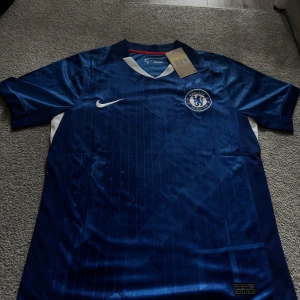 Chelsea Hemma ställ 25/26  - Chelsea FC matchtröja från Nike i Dri-Fit material. Djup blå färg med tunna vertikala ränder, vit Nike-logga och Chelsea-emblem på bröstet. V-ringad krage med vit och röd detalj, samt vita och röda inslag på sidorna. Perfekt för dig som älskar fotboll och vill representera ditt lag.