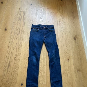 Mörkblå jeans från Nudie Jeans Co - Snygga mörkblå jeans från Nudie Jeans Co med klassisk femficksdesign och kontrastsömmar i orange. Modellen har rak passform och normal midja, tillverkade i slitstarkt bomullsjeansmaterial. Perfekt för dig som gillar stilrena och tidlösa jeans. Pris ej hugget i sten!