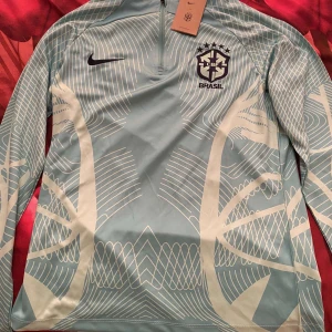 Nike Brasil dress - Snygg långärmad Brasilien fotbollströja från Nike i ljusblått med vita grafiska linjer och Brasil-logga på bröstet. Tröjan har halv dragkedja framtill och är gjord i ett lätt, sportigt material som andas. Perfekt för träning eller att representera ditt favoritlag.