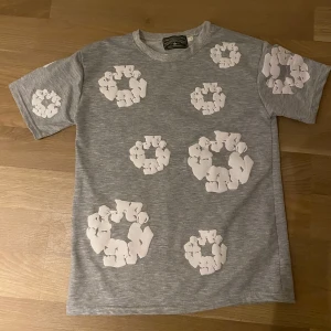 Grå t-shirt med vita tryck från Denim Tears - Säljer en grå t-shirt från Denim Tears i storlek S. T-shirten har coola vita tryck i form av cirklar över hela framsidan och på ärmarna. Klassisk rund hals och korta ärmar. Materialet är mjuk bomull som känns skön mot huden. Perfekt för dig som gillar streetwear och unik design.