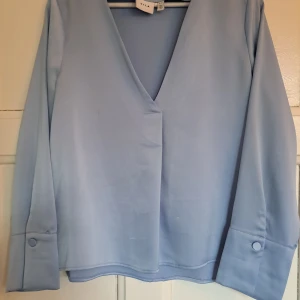 Ljusblå blus med v-ringning från Vila - Supersnygg ljusblå blus från Vila med djup v-ringning och långa ärmar. Blusen har breda manschetter med knappdetalj och en rak, lite lös passform. Perfekt att styla med jeans eller kjol för en fräsch look.