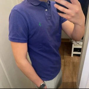 Polo Ralph Lauren (Blå piké) - Säljer en klassisk blå piké från Polo Ralph Lauren med grön broderad logga på bröstet. Pikén har krage och knappar vid halsen. Perfekt för en stilren och avslappnad look. Den är oanvänd och redo för en ny ägare! Pikén är storlek M! Slim fit.