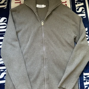  Ljung rib zip cardigan - Säljer en stilren grå ribbad kofta från Ljung med hel dragkedja och hög krage. Tröjan har långärmad design och är gjord i ett mjukt stickat material som känns skönt mot huden. Perfekt för lager-på-lager och chill dagar.