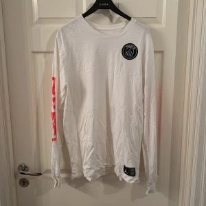 Vit PSG x Jordan långärmad tröja - Snygg vit långärmad tröja från Jordan x PSG med svart Paris-logga på bröstet och stor röd text på ena ärmen. Tillverkad i mjuk bomull, perfekt för dig som gillar sportig streetstyle. Klassisk rund hals.