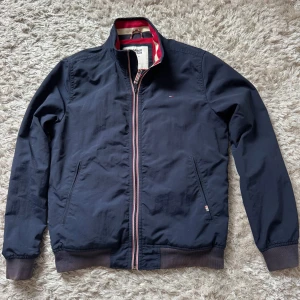 Marinblå vindjacka från Tommy Jeans - Snygg marinblå vindjacka från Tommy Jeans med klassisk ståkrage och dragkedja framtill. Jackan har ribbade muddar, två fickor och ikoniska Tommy-detaljer i rött, vitt och blått på insidan av kragen och vid dragkedjan. Perfekt för dig som gillar stilren streetwear.