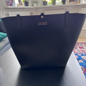 Svart axelväska från Guess - Snygg svart axelväska från Guess i strukturerat skinnimitation med guldiga detaljer och logga framtill. Väskan har två långa axelband och ett stilrent spänne upptill. Insidan är ljus och rymlig, perfekt för allt du behöver ha med dig. Nypris 1400. Mitt pris 999kr