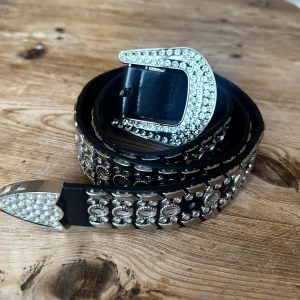 Svart glittrigt bälte med strassdetaljer - Snyggt svart bälte med massor av glittrande strass och silverfärgade detaljer. Bältet har en bred design och ett stort, dekorerat spänne som verkligen sticker ut. Perfekt accessoar för att ge outfiten extra edge och bling.  Ca 1 meter. Nypris ca 700:-