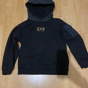 Svart EA7 hoodie med ficka på ärmen 150 cm - Svart hoodie från EA7 Emporio Armani med guldtryck på bröstet. Tröjan har en stor huva, lång ärm och en cool ficka med lock på ena ärmen. Mjuk bomullskänsla och klassisk passform, perfekt för chill dagar. Snygg och enkel design som sticker ut med loggan. Köpt för 1000