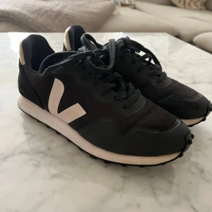 Svarta Veja sneakers med vit logga - Snygga svarta Veja sneakers med vit logga på sidan och vit detalj på hälen. Skorna har snörning, rund tå och platt sula. Ovandelen är i mesh och mocka för en sportig och clean look. Perfekta för dig som gillar stilrena och trendiga sneakers.