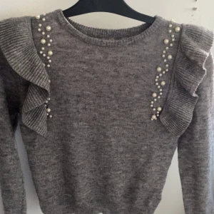 Grå stickad tröja med pärlor och volang - Säljer denna tröjan då den blivit för liten, vet ej vart är ifrån men antingen h&m eller Gina tricot. Den är super fin på 