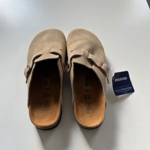 Beige mockasandaler från Birkenstock - Stilrena sandaler från Birkenstock i beige mocka med klassisk rem och metallspänne. Anatomisk fotbädd och öppen häl ger en avslappnad vibe. Perfekta för dig som gillar minimalistisk och tidlös stil. Passar mer 43