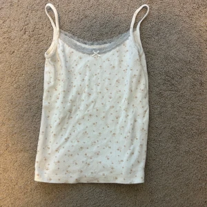 Blommig linnetopp från Brandy Melville - Supersöt vit linnetopp från Brandy Melville med små beige blommor, spetskant i grått och en liten rosett framtill. Tunt och mjukt bomullsmaterial med smala axelband.