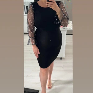 Svart kort klänning med prickiga ärmar - Snygg svart klänning med transparenta långärmade mesh-ärmar som har vita prickar och volang vid ärmslut. Klänningen är figurnära och har en rund halsringning. Perfekt för dig som gillar en stilren look med en twist.