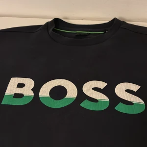 Svart BOSS sweatshirt med logga - Svart sweatshirt från BOSS i storlek S med stor vit och grön BOSS-logga framtill. Tröjan har rund halsringning och en clean, stilren look med en grön detalj på insidan av kragen.