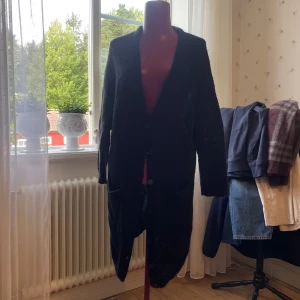 Svart lång cardigan jacka från fnk - Mysig svart lång cardigan jacka från frk med knappar framtill och stora fickor. Tillverkad i en fluffig mix av akryl, mohairull och ull som gör den varm och mysig.