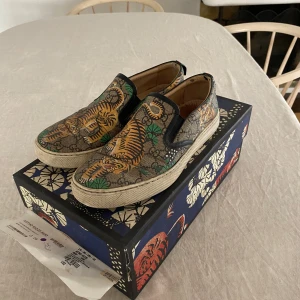 Gucci slip-on sneakers med tigerprint - Unika slip-on sneakers från Gucci med ikoniskt GG-mönster. Storlek 42,5. Nypris ca 6000kr. Orginalkvitto finns. Har varit lat och inte orkat tvätta skorna, går att fräscha upp rätt rejält!
