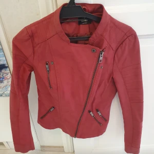 Röd skinnjacka från ONLY, stl 34 - Snygg röd skinnjacka från ONLY med coola detaljer som sned dragkedja och flera fickor med dragkedjor. Jackan har lång ärm, markerade axlar och en figurnära passform. Perfekt för dig som vill sticka ut med en färgstark look.