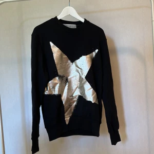 Svart tröja med silverstjärna Golden Goose - Cool svart tröja från Golden Goose med en stor, glittrig silverstjärna på framsidan. Rund hals och långa ärmar, tillverkad i mjuk bomull. Snygg logga baktill i silver.