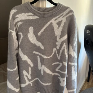 Grå stickad tröja med vitt mönster - Säljer en grå stickad tröja med vitt abstrakt mönster, tröjan har rund hals och långa ärmar. Aldrig använd pga beställd fel storlek