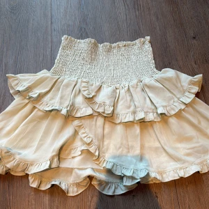 Volangkjol från Neo Noir i ljus beige - Supersöt kort volangkjol från Neo Noir i ljus beige. Kjolen har bred smock i midjan och flera lager med volanger som ger en fluffig och trendig look. Perfekt att styla med en croppad topp eller stickad tröja. Materialet känns mjukt och lätt.