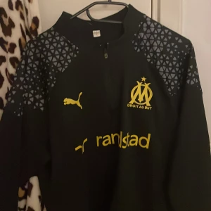 Svart långärmad Marseille Puma tröja  M/S BYXOR TILL SETTET INGÅR - Snygg svart långärmad fotbollströja från Puma med Olympique de Marseille-logga i gult på bröstet. Tröjan har gul Puma-logga och sponsortryck, samt coolt geometriskt mönster på axlarna. Materialet är mjukt och sportigt, perfekt för träning eller match.