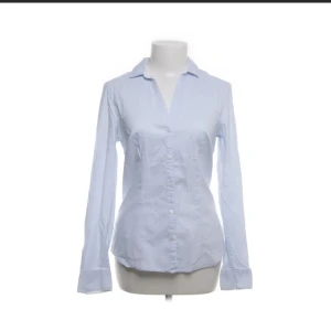 Ljusblå skjorta från H&M, strl 40 - Snygg ljusblå randig skjorta från H&M i storlek 40. Klassisk modell med krage, långa ärmar och knappar framtill. Skjortan är tillverkad i mjuk bomull och har en figurnära passform som ger en clean och fräsch look. Perfekt till jeans eller kostymbyxor. Liten i storleken så skulle säga mer 36-38