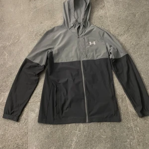 Grå och svart vindjacka från Under Armour - Säljer en snygg tvåfärgad vindjacka från Under Armour i grått och svart. Jackan har huva, dragkedja framtill, långa ärmar och logga på bröstet. Perfekt för blåsiga dagar och sportiga outfits. Knappt använd, inga hål,inga trasiga sömmar eller fläckar.