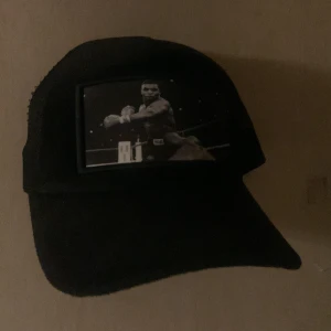 Svart velvet keps - Säljer en svart keps med mesh på sidorna och ett stort tryck framtill som visar en boxare i ringen. Skärmen är böjd och kepsen har klassisk trucker-stil. Perfekt för dig som gillar sportiga och coola accessoarer.