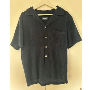 Blå frottéskjorta från Nikben - Blå kortärmad skjorta från Nikben i mjuk frotté. Klassisk krage, knappstängning framtill och en bröstficka. Perfekt för chill dagar eller när du vill ha något bekvämt och stilrent. Enkel och clean look med loose passform.