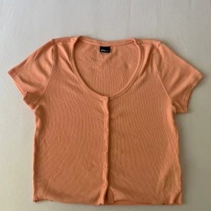 Persikofärgad ribbad topp Gina Tricot - Kortärmad, croppad topp i persikofärg från Gina Tricot. Toppen har ribbad struktur, rundad urringning och knappar framtill. Tillverkad i mjuk bomull från deras good collection. Perfekt för en chill och trendig look.