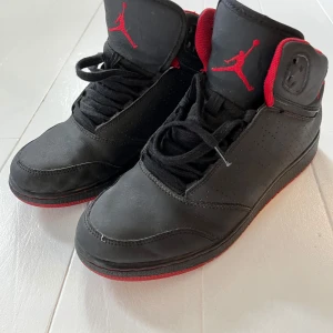 Svarta Nike Jordan 1 Highs strl 39 - Säljer ett par svarta Nike Jordan 1 Highs med röda detaljer och röd sula. Skorna har klassisk snörning, perforerade sidor och den ikoniska Jumpman-loggan på plösen. Insidan är röd och bak på hälen finns siffrorna 2 och 3. Perfekt för dig som gillar streetstyle.