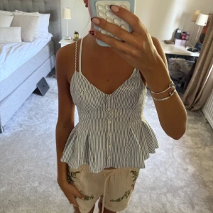 Randig vit och blå linnetopp med knappar - Supersöt linnetopp med smala axelband och v-ringad skärning. Toppen har vertikala blå och vita ränder, knappar framtill och en volangig peplum-nederdel som ger en snygg siluett. 