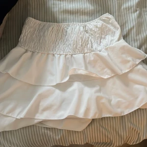 Vit volangkjol med smock - Supersöt vit kortkjol med bred smock i midjan och dubbla volanger. Kjolen har en luftig och flowy känsla, perfekt för varma dagar. Passar dig som gillar romantisk och feminin stil.