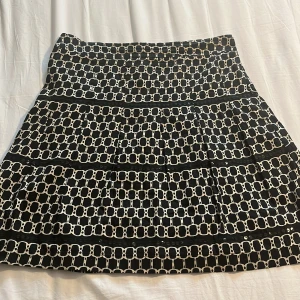 Svartvit mönstrad kortkjol - Säljer en svartvit kortkjol med grafiskt rutigt mönster och dekorativa knappar längs vissa ränder. Kjolen har en lätt utsvängd form och är tillverkad i ett mjukt, tunt material som känns skönt mot huden. Perfekt för dig som gillar statement-plagg med coola detaljer.